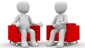 Zwei Figuren sitzen sich auf Sesseln gegenüber.